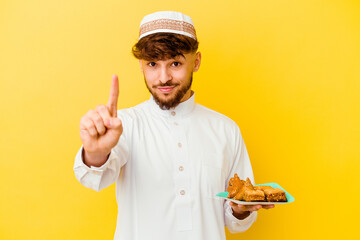 Young Moroccan man wearing the typical arabic costume eating Arabian sweets isolated on yellow background showing number one with finger.