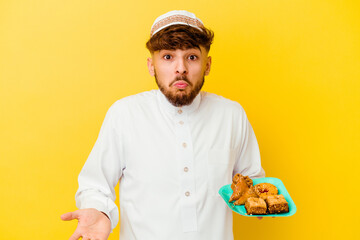 Young Moroccan man wearing the typical arabic costume eating Arabian sweets isolated on yellow background shrugs shoulders and open eyes confused.