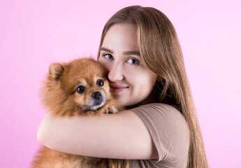 Beautiful redhead young woman hugs and kisses her puppy spitz dog. Love between dog and owner. Isolated on a pink background.