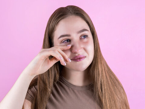 Close-up portrait of young beautiful woman crying isolated on pink, wipes tears, looking at camera
