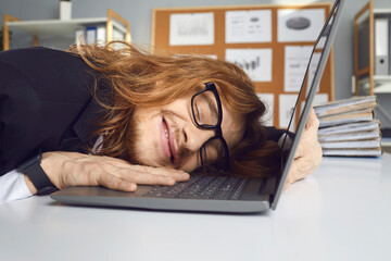 Happy office worker who loves his job tenderly hugging laptop and smiling with eyes closed. Funny young man taking nap at work, lying on computer keyboard and dreaming sweet dreams during workday
