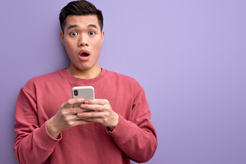 emotional asian guy chatting with someone on smartphone, stand in shock by news, surprised by message, reacts on something looking at camera. isolated purple background