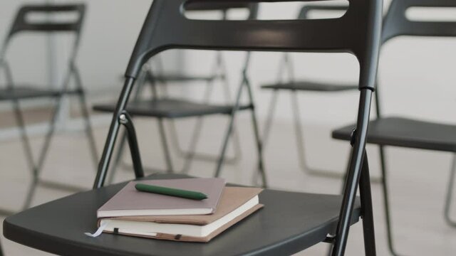 Tracking left shot of black folding chairs set up in conference room for event, notebooks and pen on one of them