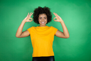 African american woman wearing orange casual shirt over green background doing ok sign with fingers and smiling, excellent symbol