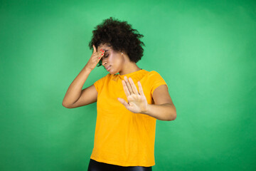 African american woman wearing orange casual shirt over green background covering eyes with hands and doing stop gesture with sad and fear expression. Embarrassed and negative concept.