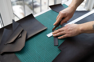 Men's hand holding a stationery knife and metal ruler and cutting on a pieces for a leather wallet in his workshop. Working process with a brown natural leather. Craftsman holding a crafting tools.