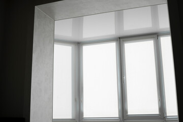 White fabric roller blinds on the plastic window on a balcony in the living room with a reflection in a stretch ceiling.