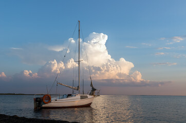 A small yacht docked by the coast against an evening sky with big cloud. Sunset over Black sea, summer time. Family vacation or travel by sea on own yacht.