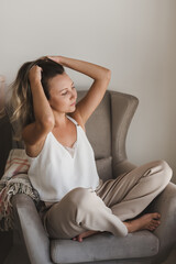 Young caucasian woman in white blouse and grey pants sitting in a comfy armchair and doing self scalp massage. Loungewear fashion. Home coziness and comfort - 419122138