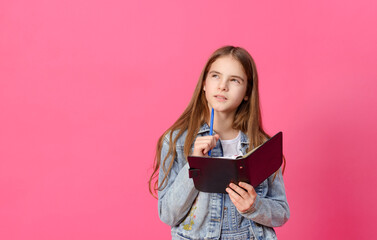  pensive white girl 10 years old in a blue denim jacket with a notebook and pen on a pink background