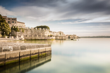 Fototapeta premium Ortigia, Syracuse, Italy / December 2018: Coast of Ortigia island at city of Syracuse. Long exposure sea. Reflection in water. Landscape reflection in sea