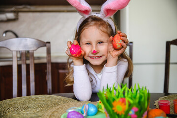 Smiling little brunette girl with bunny ears and painted nose sitting at home at the table holding painted Easter eggs