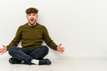 Young Moroccan man sitting on the floor isolated on white background shouting very angry, rage concept, frustrated.