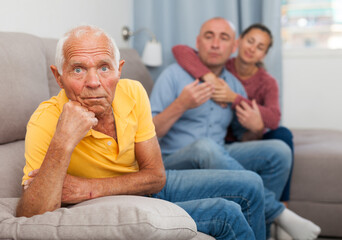 Upset senior man sitting at home on comfortable sofa, adult children on background