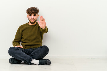 Young Moroccan man sitting on the floor isolated on white background standing with outstretched hand showing stop sign, preventing you.