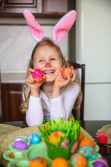 Smiling little girl with bunny ears and painted nose sitting at home at the table holding painted Easter eggs