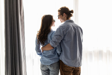 Back view of young couple looking out the window and hugging while having a breakfast at home