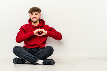 Young Moroccan man sitting on the floor isolated on white background smiling and showing a heart shape with hands.
