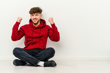 Young Moroccan man sitting on the floor isolated on white background cheering carefree and excited. Victory concept.