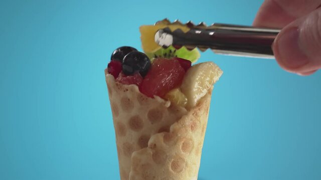 Hand puts orange wedge and blueberries onto fruit salad with tongs. Mix of fruits with blueberries in a waffle cone on a blue background. Vegan food concept.
