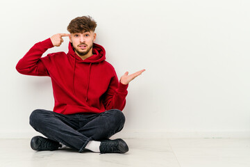 Young Moroccan man sitting on the floor isolated on white background holding and showing a product on hand.