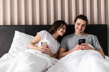Fototapeta premium Shocked woman noticing her husband checking his smartphone and texting his lover while lying next to his wife in bed. Surprised lady caught her man cheating on mobile, chatting with his mistress