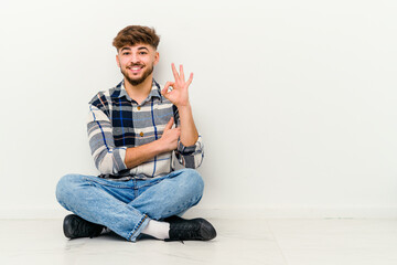 Young Moroccan man sitting on the floor isolated on white background winks an eye and holds an okay gesture with hand.