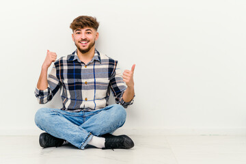 Young Moroccan man sitting on the floor isolated on white background raising both thumbs up, smiling and confident.