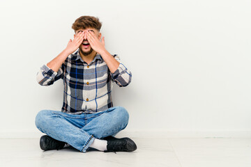 Young Moroccan man sitting on the floor isolated on white background covers eyes with hands, smiles broadly waiting for a surprise.