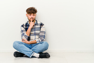 Young Moroccan man sitting on the floor isolated on white background looking sideways with doubtful and skeptical expression.