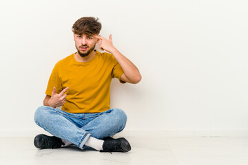 Young Moroccan man sitting on the floor isolated on white background showing a disappointment gesture with forefinger.