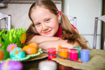 Obraz na płótnie Canvas A girl with long blond ponytails and a painted nose sits at the kitchen table in front of decorated Easter eggs