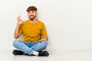 Young Moroccan man sitting on the floor isolated on white background cheerful and confident showing ok gesture.