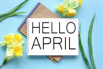 Hello April . text on white notepad paper on blue background. near notepad with yellow flowers and green leaves © Svetlana