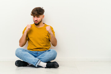 Young Moroccan man sitting on the floor isolated on white background showing fist to camera, aggressive facial expression.