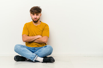 Young Moroccan man sitting on the floor isolated on white background blows cheeks, has tired expression. Facial expression concept.