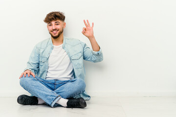 Young Moroccan man sitting on the floor isolated on white background winks an eye and holds an okay gesture with hand.