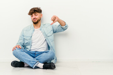 Young Moroccan man sitting on the floor isolated on white background showing a dislike gesture, thumbs down. Disagreement concept.