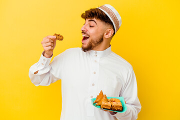 Young Moroccan man wearing the typical arabic costume eating Arabian sweets isolated on yellow background