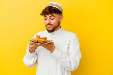 Young Moroccan man wearing the typical arabic costume eating Arabian sweets isolated on yellow background