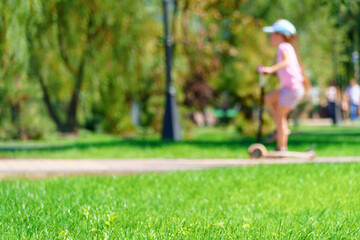 a child girl rides a scooter in a city park, summer day, green lawn with grass and trees, bright sunlight and shadows