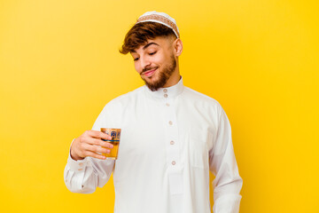 Young Moroccan man wearing the typical arabic costume drinking tea isolated on yellow background