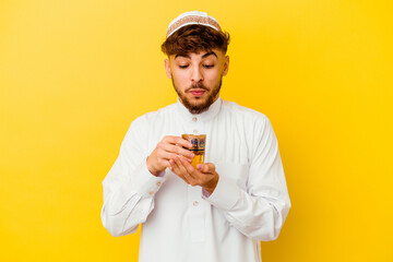 Young Moroccan man wearing the typical arabic costume drinking tea isolated on yellow background