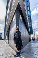 Cool girl in a protective mask in a black leather jacket posing in front of a wall  - new normal