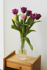 a bouquet of purple, pink and yellow tulips stands in a transparent glass vase on a wooden cabinet or table against a white wall. natural soft light. classic interior design