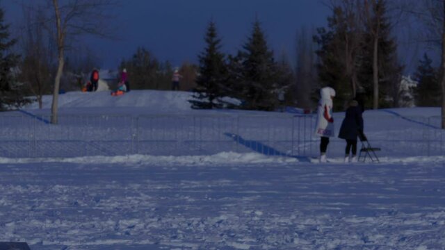Sunny ice skating lessons at park with 2 young adult ladies and one of them is leaning on a fold out chair while child wearing a helmet zips by respectfully at snowy winter park sparce pine trees etc
