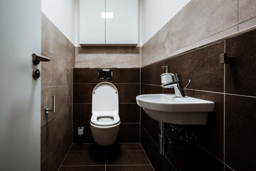 Small modern restroom with all furnishings, including a small sink and  cabinet mounted on the wall. Paving and tiling have texture of concrete trowel in shade of brown color. The toilet seat is open.
