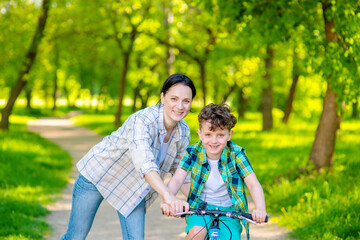 Mom helping her son learn to ride a bike in a summer park.