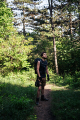 Obraz premium Man standing in forest and soaking up atmosphere. Man is dressed in shorts,undershirt and high boots.He carries large backpack with hydration bag on his back and thigh panel with equipment on his leg.