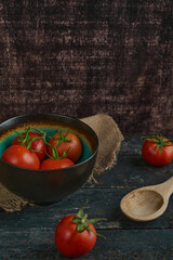 green bowl with red tomatoes, on orange cloth and dark rustic wood, with wooden spoon                             